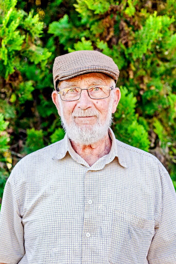 Elderly man wearing glasses and a flat cap, smiling in front of green foliage.